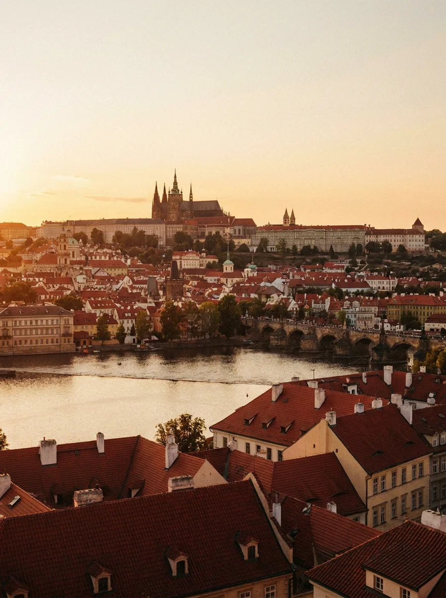 Prague at golden hour — the red rooftops of the Old Town, the Vltava river and Charles Bridge, with Prague Castle and St. Vitus Cathedral on the hill